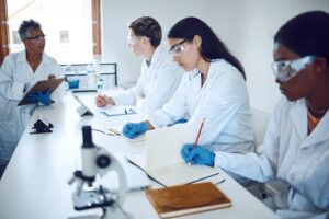 african american female professor instructs male and female students in lab coats, gloves, and safety glasses at a lab bench taking notes for academic research.